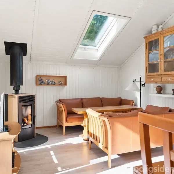 En hyggelig stue med en tændt brændeovn til venstre. Der er en hjørnesofa af træ og læder. Et glasloftovenlys giver naturligt lys. En træreol hænger på væggen.