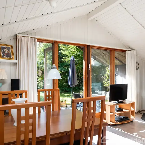 Spisebord med træstole står i en lys stue med store vinduer mod en have. En brændeovn er tændt ved væggen, og der er en tv-skærm på et bord.