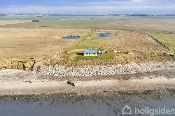 Et lille hus står alene ved en strand, omgivet af græsklædte marker og små damme. Vindmøller ses i horisonten under en blå himmel.