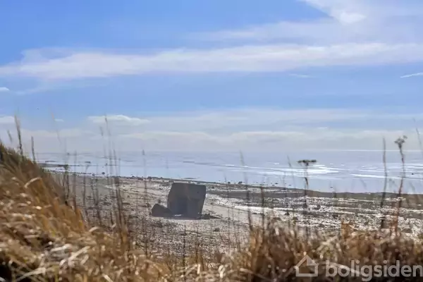 En betonbunker står stille på en sandstrand med tørre græsser i forgrunden, omgivet af hav og blå himmel.