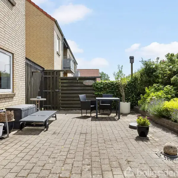 Terrasse med flisebelægning, indeholdende en liggestol, potteplanter og et bord med stole. Omgivet af en gul murstensbygning til venstre og grønne buske til højre.