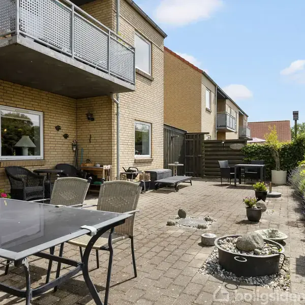 En terrasse med havemøbler og stenbede strækker sig langs en række rækkehuse. Omgivet af buske og planter, er der en rolig og solrig atmosfære.