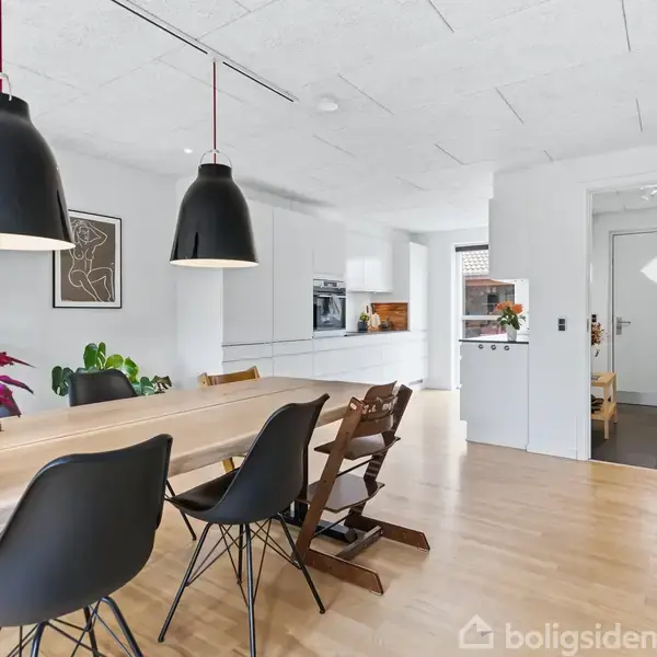Spisebord med sorte stole står under to store sorte lamper. En stor plante pryder bordet. Køkkenet ses i baggrunden, omgivet af lyse træ- og hvidelementer.