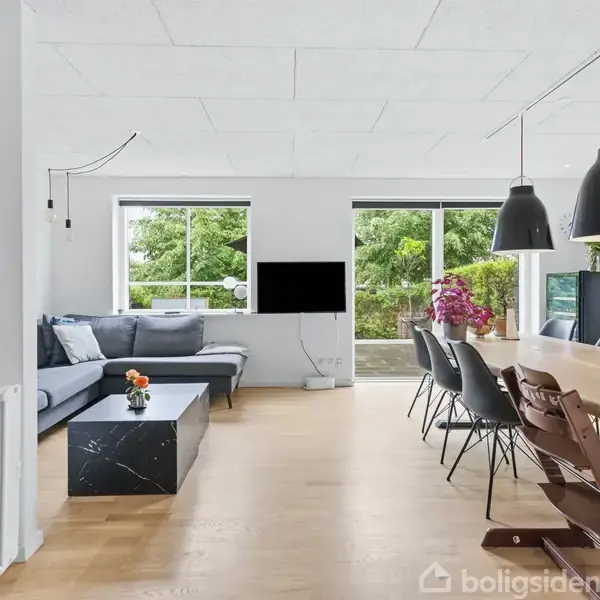 Stue med grå sofa og sofabord til venstre. Langt spisebord med stole til højre. Store vinduer bringer lys ind, med udsigt til grøn have. Moderne og lys indretning.