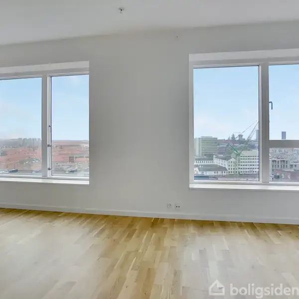 To store vinduer med udsigt til bylandskab. Rummet har lyst trægulv og hvide vægge, uden møbler. Udsigten viser bygninger og en kran i baggrunden.