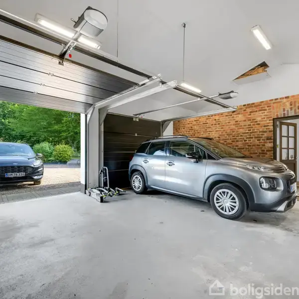 Bil holder parkeret i en rummelig garage med åben port; en anden bil står udenfor på en indkørsel. Garagen har en murstensvæg og hvidt loft.