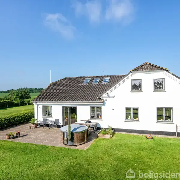 Hvidt to-etagers hus med skråt tag og store vinduer, placeret i en frodig have. Rundt badekar på terrasse og grønne marker i baggrunden under en blå himmel.