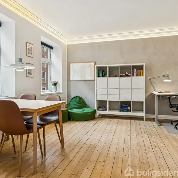 Spisebord med stole står ved siden af to vinduer; en reol og skrivebord med lampe og kontorstol står op ad en væg i et lyst rum med trægulve.
