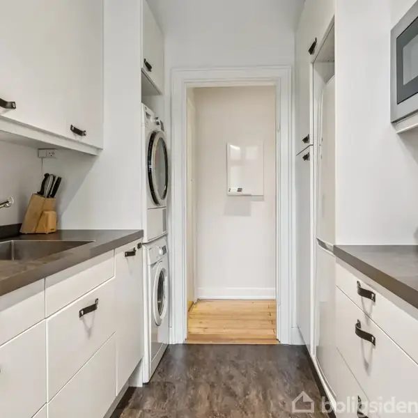 Et køkken med hvide skabe og mørke bordplader. En vask står til venstre, mens en mikroovn og en frugtskål med appelsiner er til højre. En vaskemaskine og tørretumbler er indbygget i væggen. En åben dør fører til en lysere gang.