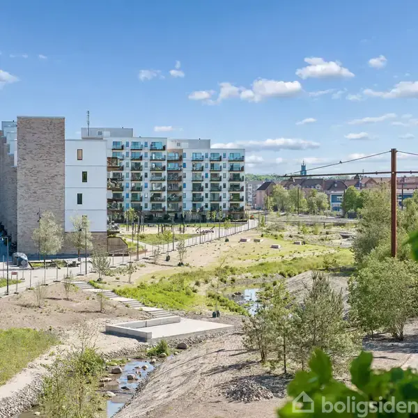Et moderne lejlighedskompleks med flere etager står ved siden af et åbent, grønt område med stier og små vandløb. Solrig dag med blå himmel og spredte skyer.