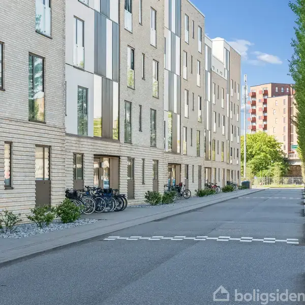 Lejlighedsbygning strækker sig langs en stille vej, hvor cykler er parkeret foran indgangen. Træer står til højre, og flere lejligheder ses i baggrunden.