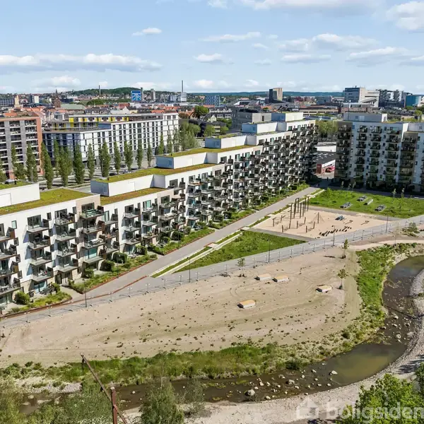 Flere moderne lejlighedsbygninger strækker sig langs et landskab med græs og sand, omfavnet af en snoet flod. Byens skyline kan ses i baggrunden under en blå himmel.