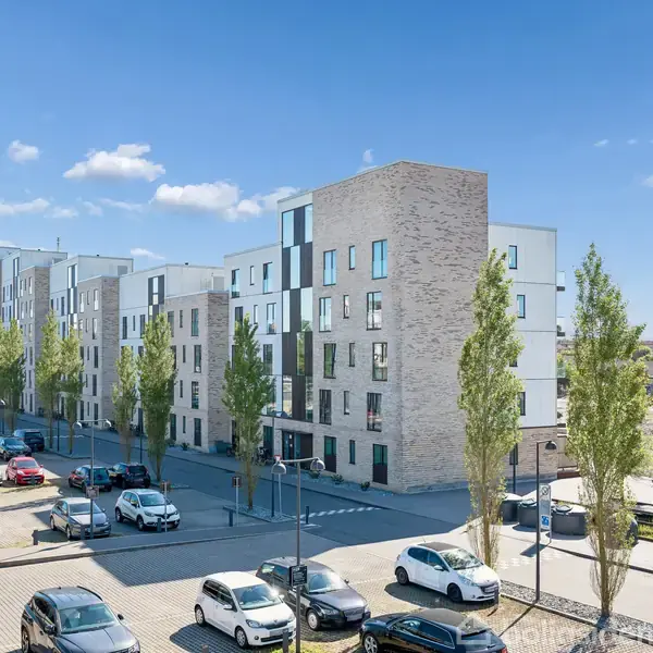 Bygning i fem etager står stille i sollys, omgivet af parkerede biler og træer. Klar blå himmel og spredte skyer dominerer baggrunden.