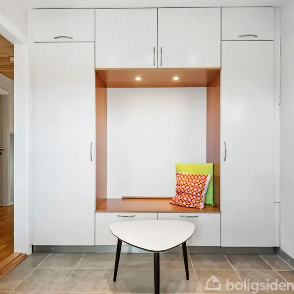 Et hvidt skabssystem fylder væggen og har en indbygget bænk med farverige puder. Foran bænken står et lille hvidt bord. Rummet er lyst og ryddeligt.