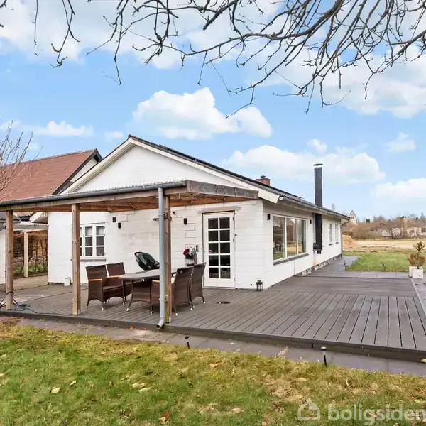 Et hvidt hus står med en overdækket terrasse, hvor et bord og stole er placeret. Omgivet af en græsplæne og nøgne træer under en blå himmel med skyer.