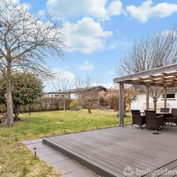 Træterrasse med møbler står under en pergola i en have. Græsplæne og træer omgiver området, mens en skyfri blå himmel ses i baggrunden.