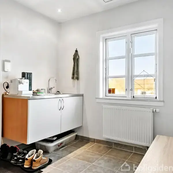 En entre med sko placeret ved en dør, en skænk med håndvask og køkkenredskaber til venstre, og et vindue og en radiator på højre væg.