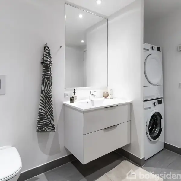 Et moderne badeværelse med en vaskemaskine og tørretumbler stablet til højre. En hvid vask med spejl hænger på væggen, omgivet af lyse grå fliser og et toilet.