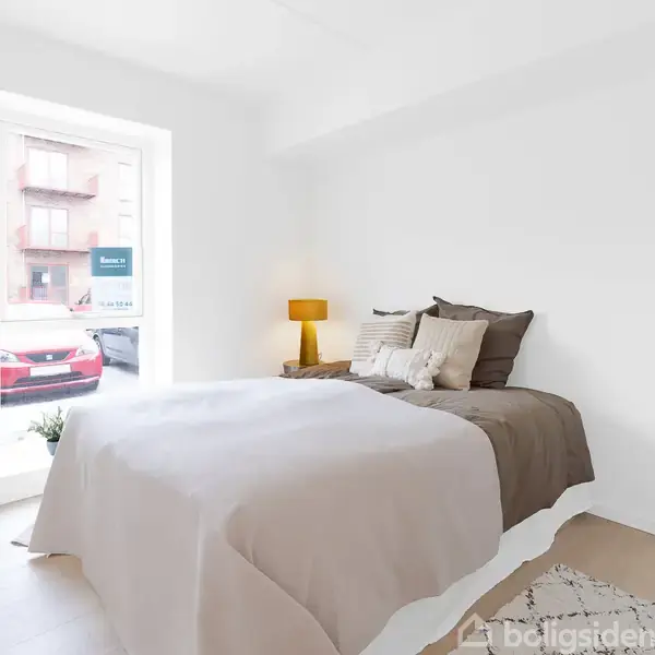 En dobbeltseng med grå og beige sengetøj står i et lyst soveværelse. Ved siden af sengen er en orange lampe og en grøn plante ved vinduet. Udenfor ses en parkeret bil.
