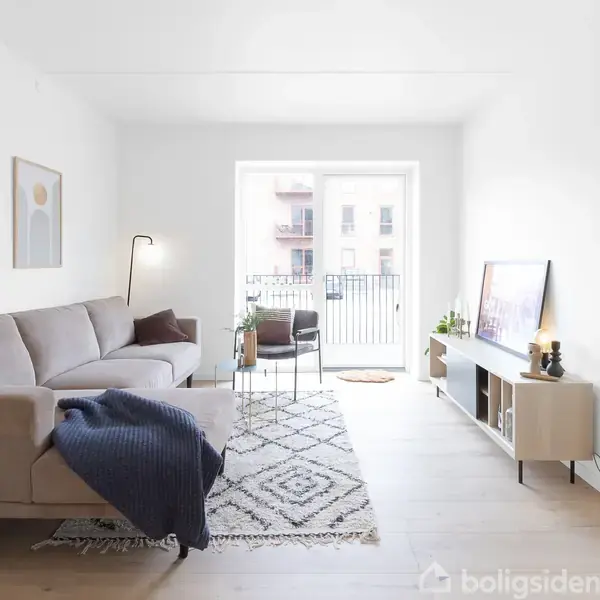 Sofa med blå tæppe placeret i stue; gulvlampe ved siden af; lysindfald fra terrassedør bagved; vægdekorationer og TV på skab til højre.