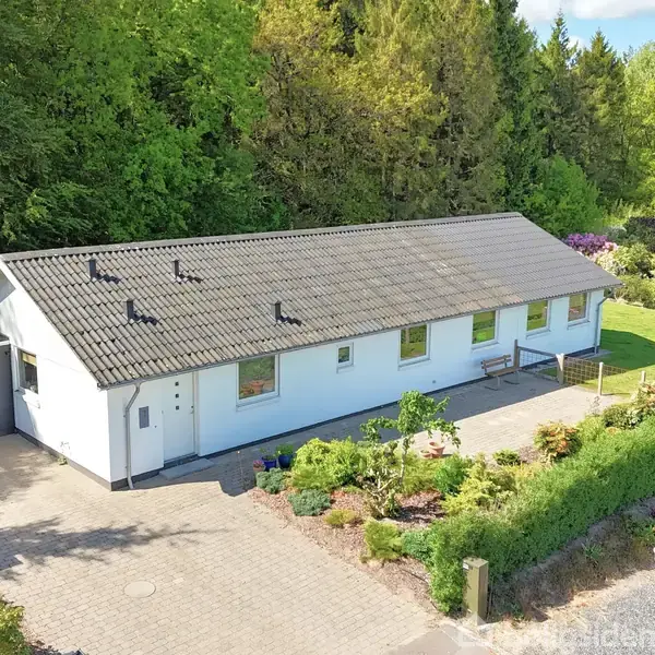 Hvidt hus med gråt tag ligger i en frodig have med stier og bede. Foran huset er der en indkørsel og carport. Omkring huset er der grønne træer og buske.