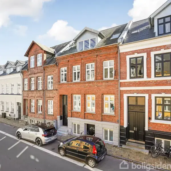 Rækkehuse i forskellig farvet mursten står på en byvej. Foran husene er parkerede biler og cykler. Himlen er klar med nogle få skyer.