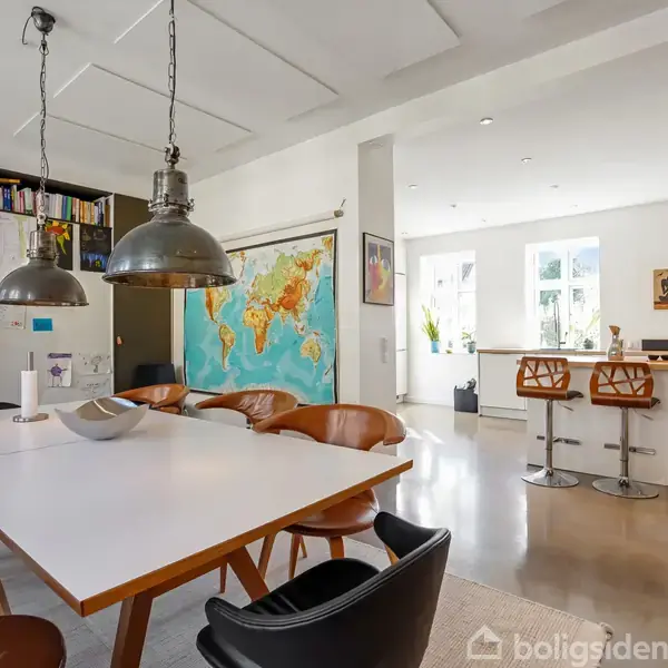 Et spisebord med stole omkring står i et moderne køkken-alrum. Kort på væggen, industrielle loftslamper og en ø-bar med høje stole. Lyse vægge og gulv.
