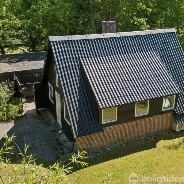 Et sort tagklædt hus med røde murstensvægge står i en frodig have omgivet af grønne træer. En skyggefuld terrasse ses ved siden af huset.