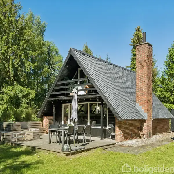 A-formet sommerhus med sort tag og murstensskorsten, står på en terrasse med havemøbler, omgivet af en grøn græsplæne og skov. Skyderutsjebane ses til venstre.
