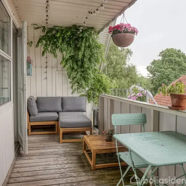 Balkon med træmøbler og et gråt sofaarrangement. Blomster og grønne planter hænger fra taget og står på kanten. Omringet af træer og udsigt til hustage.