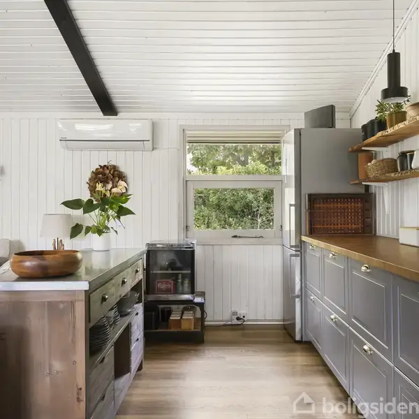 Køkken med grå skabe og en træbordplade. Hylder over skabene har forskellige krukker og kopper. Et vindue viser grønne træer udenfor. En stor plante står ved en grå sofa.