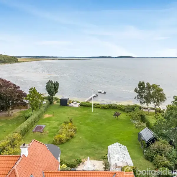 En stor, velholdt have med grønne plæner og enkelte træer strækker sig ned til en bred fjord med en lille bådebro. Huset har rødt tegltag og er omgivet af frodig vegetation.