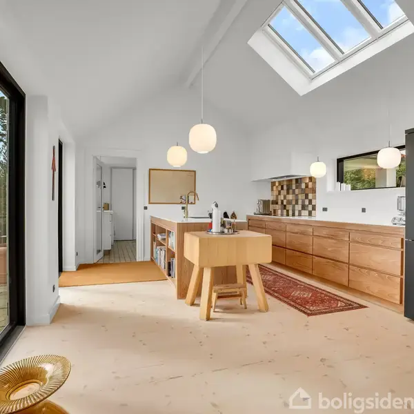 Køkken med lyst træbord, hængende lamper og moderne hvide elementer. Rummet har store vinduer, en sort skydedør og et gulvtæppe, omgivet af lyse vægge i et åbent rum.