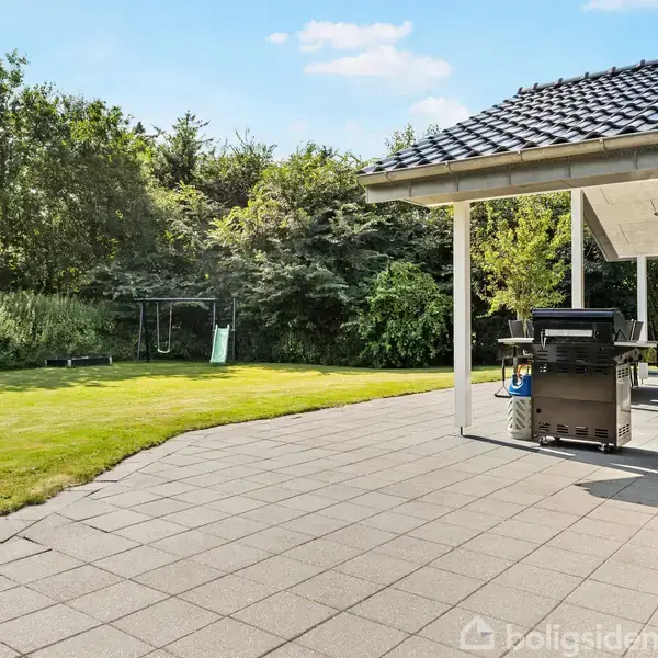 Terrasse med havemøbler og grill ved siden af et hus. Græsplæne og gyngestativ i baggrunden omgivet af buske og træer.