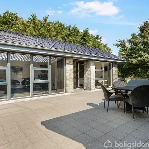 Et moderne hus med store vinduesfacader står på en flisebelagt terrasse. Et spisebord med stole er placeret tæt på huset. Baggrunden prydes af grønne træer og blå himmel.
