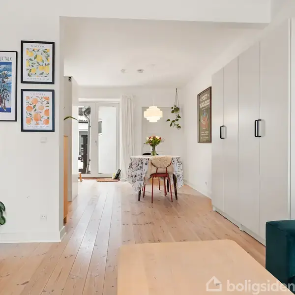 Sofa står mod væg med puder og tæpper, overfor et træbord. Fjernsyn hænger på væggen. Rummet har lyse trægulve, grønne planter og store vinduer med dagslys.