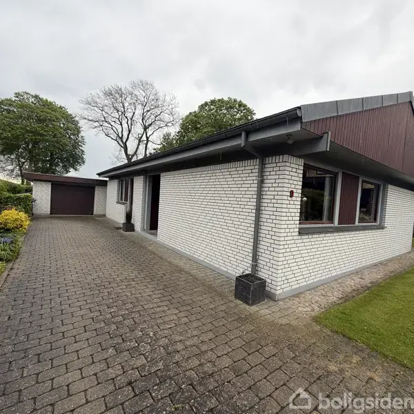 En hvid murstensvilla står stille ved siden af en flisebelagt indkørsel, der fører til en garage. Haven omkranser huset med træer og buske i baggrunden.