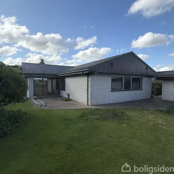 En moderne, hvid murstensejendom med skrånende tag står stille i en velplejet have. Baggrunden viser en blå himmel med skyer.