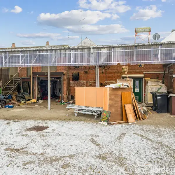 En gårdsplads med en lang, gammel murstensbygning bagved. Bygningen har en tagkonstruktion af gennemsigtigt materiale med diverse opmagasinerede materialer, træplader og værktøj spredt rundt i området.