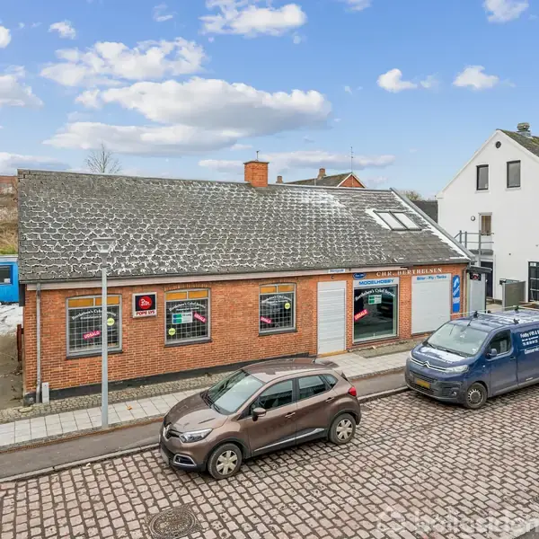 En murstensbygning med skråt tag står langs en brostensbelagt vej. Foran bygningen holder en brun bil og to blå varevogne parkeret. Himlen er skyet med spredte skyer.