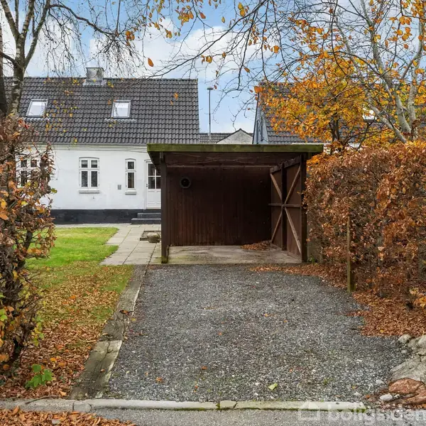 Et brunt skur står tomt ved siden af et grusbelagt område. Omkranset af efterårsløv og grønne plæner ligger et hvidt hus bagved.