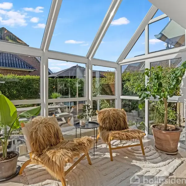 To lænestole med pelssæder står i en solrig udestue omgivet af store planter. Store vinduer viser en udsigt til en have og lyseblå himmel med skyer.