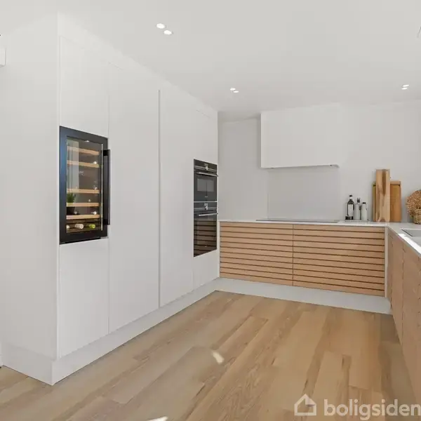 Hvidt og træfarvet køkken med integrerede apparater, herunder en ovn og vinkøleskab, på højre væg. Rummeligt med træparketgulv og et stort vindue over køkkenbordet.