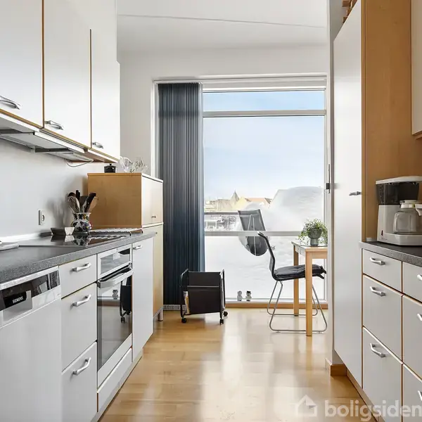 Et moderne køkken med hvide skabe og apparater; der er en kaffemaskine og en ovn. En lille spiseplads med stole og et vindue viser udsigt til en by.