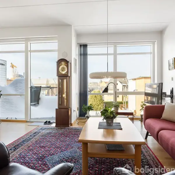 Stue med rødt tæppe og rød sofa, sofabord med planter, pendellampe hænger over bordet, store vinduer med tynde gardiner, udsigt til by samt et spisebord ved siden af skabet.