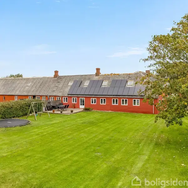 Rødt, langstrakt hus med solpaneler på taget står i en rummelig grøn have. Foran huset er der en trampolin og en gyngestativ i et åbent område med græsplæne.