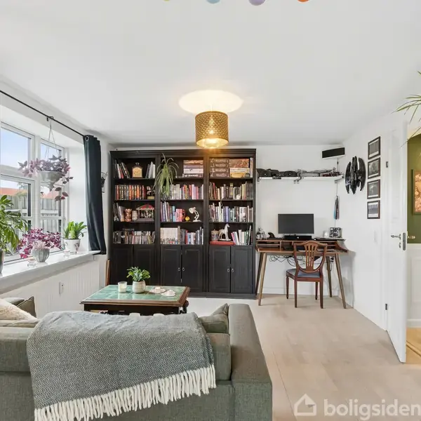 En lys stue med en grøn sofa, hvor et gråt tæppe ligger henover ryglænet. Der er en reol fyldt med bøger. Et skrivebord med computer står ved siden af. Store vinduer med planter giver naturligt lys. En åben dør fører til et andet rum.