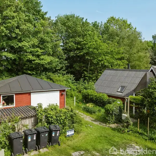 To huse af træ, begge med skrå tage, står omringet af frodig vegetation. Fire skraldespande står langs en sti nær huse, omgivet af grønne træer og buske i landskabet.