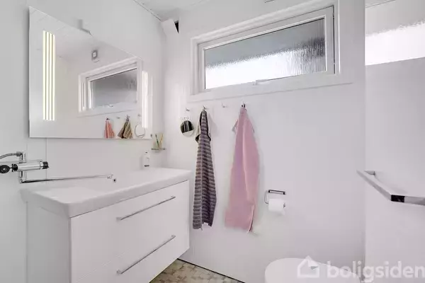 Et moderne badeværelse med en hvid vask og skab. Tre håndklæder hænger på kroge under et vindue. Rummet har et stort spejl og fliser på gulvet.