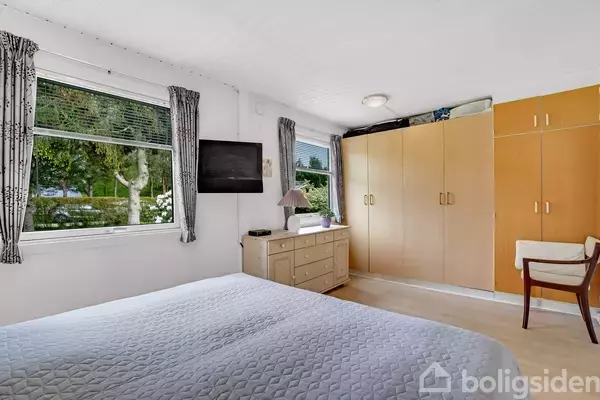 Soveværelse med dobbeltseng, klædeskab og kommode med lampe. Et vægmonteret TV hænger ved siden af et vindue med udsigt til en grøn have. Gardiner rammer vinduerne.